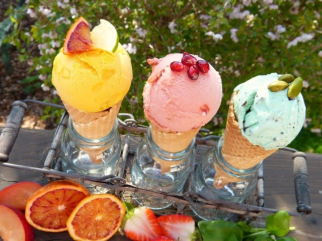 zmrzlina tři vedle sebe, žlutý kopeček zmrzliny, vedle je červená a pak zelená, za nimi je keř, stojí ve skleněných nádobách, kolem jsou jahody, pomeranče 